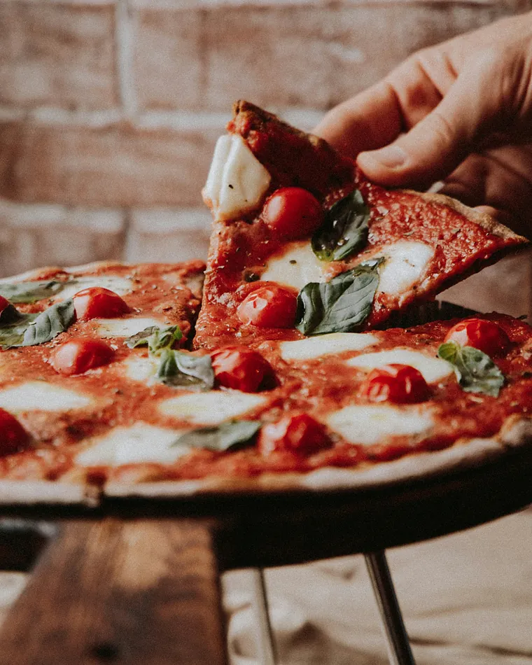 Una mano solleva uno spicchio di pizza con pomodorini, mozzarella e basilico (immagine illustrativa)